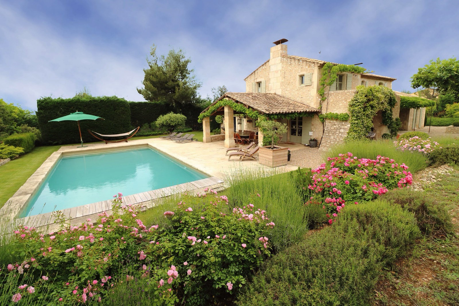 A luxurious stone villa with a swimming pool in the foreground, surrounded by a lush garden with vibrant pink flowers, under a clear blue sky.